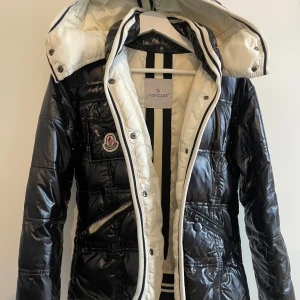 Moncler dunjacka - Snygg svart glansig dunjacka från Moncler med vit insida och avtagbar huva. Jackan har dragkedja, knappar, flera fickor och klassisk Moncler-logga på bröstet. I använt skick men inget trasigt/defekt. 