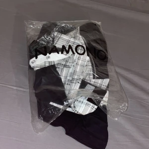 Svart och rutig henley från Namono - Cool långärmad henley från Namono med svart bas och rutigt vitt och grått tyg på krage och axlar. Snyggt tryck framtill och detaljerade knappar vid halsen. Perfekt för dig som gillar unika skjortor med streetkänsla.