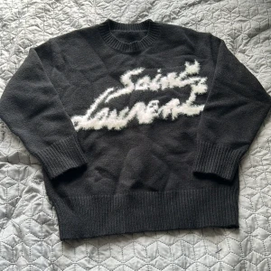 Svart stickad Saint Laurent tröja - Säljer en svart stickad tröja från Saint Laurent med vit, fluffig logotyp framtill. Tröjan har rund hals, långa ärmar och breda ribbade muddar vid ärmslut och nederkant. Perfekt för dig som gillar streetstyle och exklusiva märken.