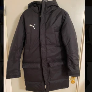 TeamFINAL vinterjacka Puma - Säljer en svart lång pufferjacka från Puma med huva och dragkedja framtill. Jackan har två stora fickor med lock och Puma-logga på bröstet. Perfekt för kalla dagar när du vill hålla dig varm och ändå se stilren ut.