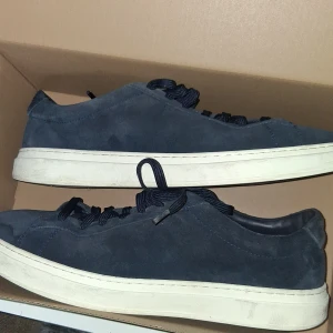 Mörkblå mockasneakers från BOSS - Stilrena mörkblå sneakers från BOSS i mjuk mocka med vita sulor och svarta snören. Klassisk låg modell med diskret BOSS-logga på sidan. Perfekta för dig som gillar en clean och modern look. Köptes för 2099 i nk varuhus finns bilder på det. Nästan aldrig använts köptes för 1 månad sedan