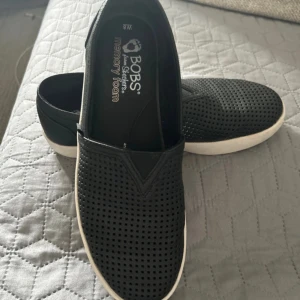 Svarta slip-on sneakers från BOBS - Svarta slip-on sneakers från BOBS by Skechers med vit sula och perforerat mönster på ovandelen. Skorna har memory foam-innersula för extra komfort och en klassisk, rund tå. Perfekta för en avslappnad och stilren look.