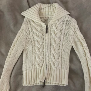 Vit stickad kofta från Mng med dragkedja - Säljer en chunky vit stickad kofta från Mng i storlek M. Koftan har grova flätstickade detaljer, bred ribbad krage och dragkedja framtill. Perfekt för lager-på-lager och ger en mysig vibe till din outfit.