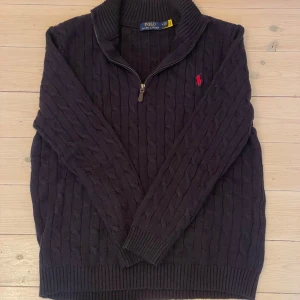 Mörkblå stickad tröja Polo Ralph Lauren - Kabelstickad ull mörkblå tröja från Polo Ralph Lauren med halv dragkedja och röd broderad logga på bröstet. Tröjan har hög krage och långa ärmar. Klassisk och stilren design som funkar till det mesta.