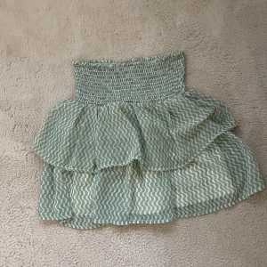 Grön mönstrad volangkjol Kappahl - Supersöt grön kortkjol från Kappahl med vågigt mönster och bred smock i midjan. Kjolen har dubbla volanger som ger en extra flowig känsla och är gjord i ett lätt och luftigt material. Perfekt för dig som gillar att sticka ut med coola detaljer och mönster. Säljer då den inte kommer till användning ❤️använt 1 gång.