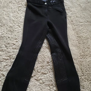 Svarta ridbyxor från CRW, stl 140 - Svarta ridbyxor från CRW i storlek 140. Byxorna har helskodd silikongrepp längs insidan av benen för extra fäste i sadeln. De har en smidig dragkedja fram och en diskret logga i midjan. Perfekta för dig som gillar ridning och vill ha bra grepp.