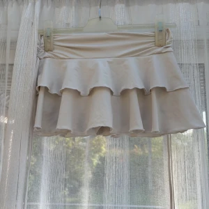 Beige volangkjol från SHEIN - Supersöt beige kortkjol från SHEIN med dubbla volanger och bred resår i midjan. Kjolen har inbyggda shorts under för extra komfort och är gjord i ett mjukt, stretchigt material. Perfekt för dig som gillar en lekfull och trendig stil.