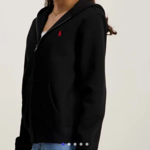 Svart hoodie från Ralph Lauren - Svart hoodie från Ralph Lauren med dragkedja och huva. Klassisk röd logga broderad på bröstet. Mjuk bomullskänsla, känguruficka framtill och långa ärmar. Perfekt till jeans eller joggers för en chill look.