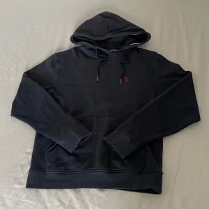 Mörkblå hoodie från U.S. Polo Assn. - Mörkblå hoodie från U.S. Polo Assn. med röd broderad logga på bröstet. Tröjan har huva med snörning, känguruficka och är tillverkad i 100% bomull. Perfekt för dig som gillar en clean och klassisk stil.