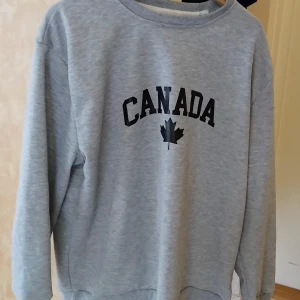 Grå sweatshirt med Canada-tryck - Säljer en grå sweatshirt med svart text 'CANADA' och ett lönnlöv på bröstet. Tröjan har rund halsringning och långa ärmar. Perfekt för dig som gillar enkel och sportig stil.