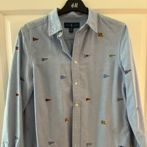 Ralph lauren stjorta  - Mycket Snygg ljusblå skjorta från Ralph Lauren med broderade små flaggor och RL-detaljer i olika färger.  Storleken är xl kids men passar S/ Nyskick!