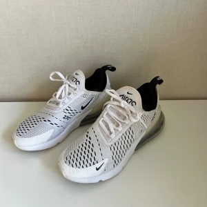 Nike Air Max 270 vita sneakers - Säljer ett par fräscha Nike Air Max 270 sneakers i vitt med svarta detaljer. Skorna har en luftig mesh-överdel, snörning och en tydlig synlig Air Max-sula baktill. Perfekta för dig som gillar sportig och modern stil.
