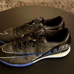 Nike Mercurial fotbollsskor svart/blå - Snygga Nike Mercurial fotbollsskor med svart ovandel, blå sula och vita detaljer. Skorna har snörning och är tillverkade i syntetmaterial för bästa känsla på planen. Perfekta för dig som vill ha stilrena och snabba skor till inomhusfotboll.