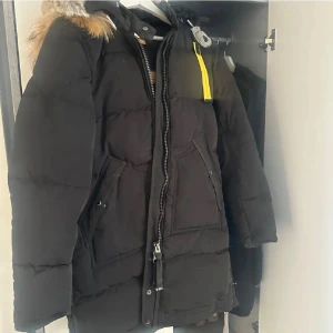 Svart Parajumpers dunjacka med päls - Säljer en svart Parajumpers Anchorage dunjacka med snygg päls på huvan och coola detaljer som gul rem och patch på ärmen. Jackan har dragkedja, stora fickor och är perfekt för kalla vinterdagar. Fodret är mjukt och jackan är riktigt varm och skön.