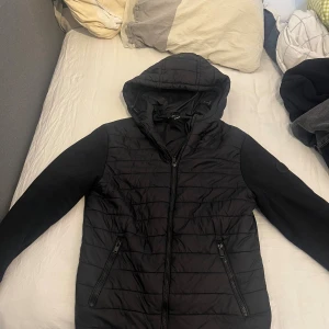 Svart cardigan från Lindbergh  - Svart cardigan från Lindbergh med huva och dragkedja framtill. Jackan har quiltad framsida, släta ärmar och två fickor med dragkedja. Perfekt för höst och vår dagar och har en clean, stilren look. Storlek M Köpt för 1799kr. Mitt pris 600kr. Tvär fet cardigan med skick 9/10. Vid fler frågor eller intresse skriv! Fler bilder kan skickas!