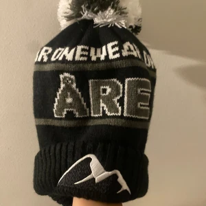 Svart och grå Åre-mössa med tofs - Stickad svart mössa från Onewear med grå och vita detaljer, stor text 'ÅRE' framtill och broderat bergmotiv. 