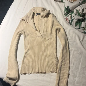 Beige ribbad tröja från Gina Tricot - Supersnygg ribbad beige tröja från Gina Tricot i figurnära modell. Tröjan har lång ärm med knappdetalj vid ärmslut, v-ringad krage och mjukt stickat material. Tröjan är i bra skick och är endast använd några få gånger! 