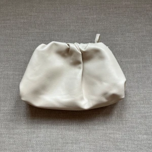 Krämvit dumpling bag  - Stilren kuvertväska i krämvitt syntetmaterial med mjuk, puffig form och rynkade detaljer upptill. Väskan har en avtagbar och justerbar axelrem, vilket gör den enkel att bära crossbody eller i handen. Perfekt för dig som gillar minimalistisk och trendig design.