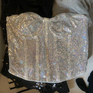 Glittrig silver korsett topp - Supersnygg korsett topp i silver med massor av glittrande paljetter. Axelbandslös bandeau-modell som verkligen fångar ljuset och ger en festlig vibe. Perfekt för dig som vill sticka ut och glänsa på nästa event!