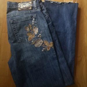 Blå jeans med broderi från Dolce & Gabbana - Snygga blå jeans från Dolce & Gabbana med unikt blommigt broderi i guld och beige på bakfickan. Klassisk femficksmodell med raka ben och mörk tvätt. Jeansen har silverfärgade nitar och tydlig D&G-logga på bakfickan. Perfekt för dig som vill sticka ut med detaljer. Avklippta. Skriv vid frågor :) 