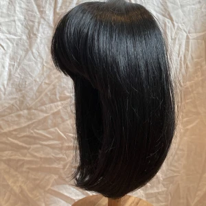 ”Cleopatra”-peruk från Rapunzel - Snygg svart peruk i bob-stil med rak lugg och glansigt, rakt hår. Perfekt för dig som vill ha en cool och edgy look. Håret är axellångt med kortklippt nacke och har en naturlig form som ramar in ansiktet. Passar till cosplay, temafest eller när du vill testa något nytt.