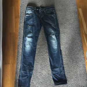 Replay Anbass blå jeans, storlek 29/32 - Säljer ett par Replay Anbass jeans i blå tvätt med slitningar och snygga detaljer. Modellen är slim fit med klassiska fem fickor och dragkedja. Jeansen är tillverkade i stretchigt denim för extra komfort och har en modern look. Slitningarna är egna!