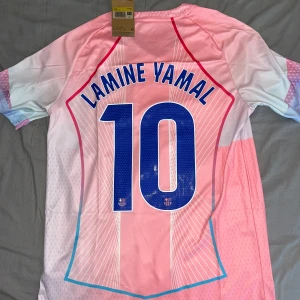 Barcelona Lamine Yamal rosa matchtröja - Säljer en FC Barcelona fotbollströja med Lamine Yamal och nummer 10 på ryggen. Tröjan är rosa med vita och blå detaljer, har korta ärmar och V-ringning. Nike-logga, klubbmärke och Spotify som sponsor framtill. Tillverkad i lätt och ventilerande material. Har 2 stycken i storlek S 