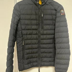 Svart dunjacka Parajumpers XS - Säljer en svart, superlätt dunjacka från Parajumpers i storlek XS. Jackan har quiltad design, dragkedjor på fickor och bröst, samt en snygg patch på ärmen. Perfekt för dig som vill ha en stilren och funktionell jacka med coola detaljer.Äkta