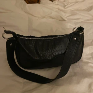 Svart axelväska - Snygg svart axelväska med croco-mönstrad yta och bred axelrem. Väskan har silverfärgad dragkedja och rundade former, perfekt för att lyfta din outfit. Materialet är syntet med struktur som påminner om krokodilskinn.