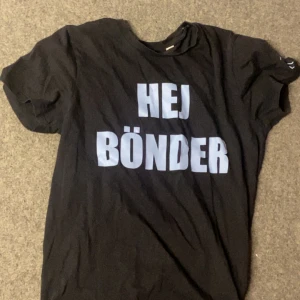 Svart t-shirt med HEJ BÖNDER-tryck - Svart t-shirt med stort vitt tryck på bröstet där det står 'HEJ BÖNDER'. Klassisk rund hals och korta ärmar. Perfekt för dig som vill sticka ut med ett roligt statement. T-shirten är gjord i mjuk bomull och har en avslappnad passform.