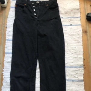 Levi's Ribcage Straight svarta jeans - Svarta Levi's Ribcage Straight jeans med hög midja och raka ben. Klassisk femficksmodell med knäppning framtill och diskreta nitar. Jeansen är i kraftigt denimtyg och har en clean look som passar till allt. Jeansen har två hål i fram som man kan se