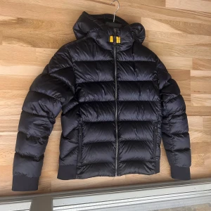 Svart dunjacka Parajumpers Sheen Y-L - Snygg svart dunjacka från Parajumpers, modell Sheen. Jackan har en puffig look, huva och dragkedja framtill. Klassiska ribbade muddar vid ärmslut och nederkant. Perfekt för kalla dagar och riktigt stilren med diskreta detaljer och gul rem vid halsen. Passar till folk mellan 165-175