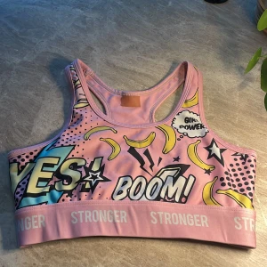 Färgglad sporttopp från Stronger - Sporttopp från Stronger i rosa med pop art-inspirerat tryck med bananer, stjärnor och text som 'YES!' och 'BOOM!'. Bred resår med logga, brottarrygg och coola detaljer. Perfekt för dig som vill sticka ut på gymmet. Utan vaddering! Säljer då jag inte använder den längre och den sitter tajt. 