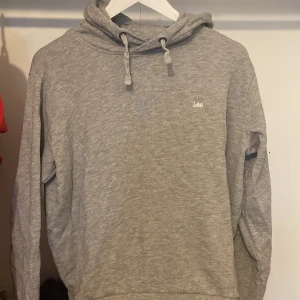 Grå hoodie från Lee med huva - Säljer en grå hoodie från Lee i regular fit med broderad logga på bröstet. Tröjan har huva med snörning och långa ärmar. Perfekt för chill dagar och enkel att matcha med jeans eller joggers.