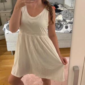 Vit kort klänning med spetsdetalj - Supersöt vit klänning med ärmlös design och rundad hals. Den har en fin spetsdetalj i midjan som ger extra charm och en luftig, flowy passform. Perfekt för varma dagar och enkel att styla med sneakers eller sandaler. Materialet känns lätt och bekvämt mot huden.
