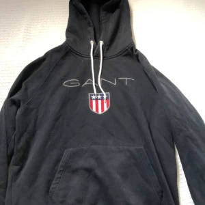 Svart hoodie från GANT med logga på framsidan - Svart hoodie från GANT i storlek 2XL med klassisk logga och sköld på bröstet. Tröjan har huva med vita snören och en stor magficka. Tillverkad i mjuk bomullsblandning (80% bomull, 20% polyester) som är perfekt för kyligare dagar.