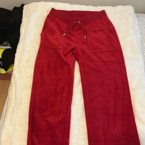 Röda velourbyxor Juicy Couture XS - Dragkedjan har gått sönder. Röda velourbyxor från Juicy Couture med bred resår i midjan och snörning. Byxorna har raka ben, stora fickor fram och bak med knappdetaljer. Perfekta för en chill och trendig look.