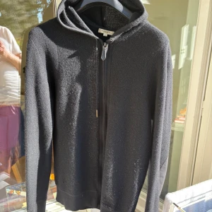 Svart hoodie från Burberry i ull - Svart hoodie från Burberry i mjuk stickad ull med dragkedja framtill och huva. Snygga mockadetaljer på armbågarna och diskret Burberry-logga på snörningen. Perfekt för dig som gillar stilren och exklusiv streetwear.