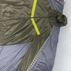 Olivgrön vindjacka från Lacoste Sport - Snygg olivgrön vindjacka från Lacoste Sport med luva och hel dragkedja. Ärmarna har mönstrad kant och materialet är lätt och syntetiskt, den använd bara några fåtal gånger, byts gärna ut med någon annan flis eller hoodie men är öppet till allt 