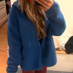 Blå fleecejacka med dragkedja - Mysig blå fleecejacka med hel dragkedja framtill och hög krage. Jackan har en relaxed passform och är perfekt för kyliga dagar. Fickor med dragkedja på bröstet och sidorna. Skön att slänga på sig över en hoodie eller t-shirt.