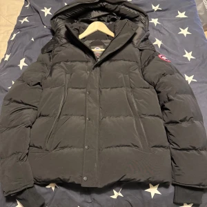 Svart dunjacka från Canada Goose  - Säljer en svart dunjacka från Canada Goose. Jackan har huva, dragkedja och knappar framtill samt klassisk logga på ärmen. Fylld med dun för extra värme och har en clean, puffig look som passar perfekt när det är kallt ute.jackan står att den är stor i storleken Skulle säga att det mer är en L passar L eller XL absolut inte större än det . Säljer för att jag vill hitta en macmillan modell så byter funkar också. Jackan är helt oanvänd så om bilder sökes går det absolut o fixa 