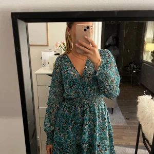 Blommig blå kortklänning med v-ringning - Säljer en superfin kortklänning med blommigt mönster i blå och gröna toner. Klänningen har v-ringad hals, långärmade transparenta ärmar och smock i midjan för en snygg passform. Materialet är lätt och luftigt, perfekt för varmare dagar. Den är lite mer grön än blå!💕