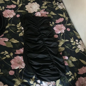 Svart kort omlottklänning med rynk - Säljer en svart kort klänning med snygga rynkade detaljer längs hela klänningen. Modellen är omlott och har en cool v-ringning. Perfekt för dig som gillar en tight och trendig look. Materialet känns mjukt och stretchigt, troligen polyester.