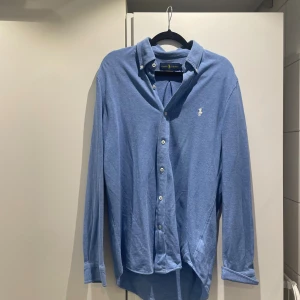 Blå långärmad skjorta från Ralph Lauren - Klassisk blå skjorta från Ralph Lauren med vit broderad logga på bröstet. Skjortan har knappar hela vägen, button down-krage och är tillverkad i mjuk bomull. Perfekt för en clean och stilren look.