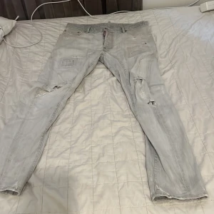 Grå slitna jeans från Dsquared2 - Säljer ett par grå jeans från Dsquared2 med coola slitningar och patchdetaljer. Jeansen har en smal passform, klassiska fem fickor och tydliga sömmar baktill. Perfekta för dig som gillar streetstyle och vill sticka ut lite extra. De är absolut äkta 