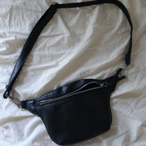 Svart magväska i skinn med dragkedja - Snygg svart magväska i skinn med justerbart axelband och silverfärgade metalldetaljer. Väskan har en stor dragkedja framtill och coola knyt-detaljer vid dragkedjan. Perfekt för att bära det viktigaste och hålla händerna fria.