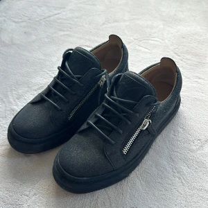 Svarta sneakers från Giuseppe Zanotti - Snygga svarta sneakers från Giuseppe Zanotti med glittrig finish och coola dragkedjor på sidorna. Skorna har snörning, rund tå och platt sula. Utsidan är i mocka och insidan i skinn för extra komfort. Perfekta för dig som gillar statement-detaljer. Kvitto skickas på begäran. 