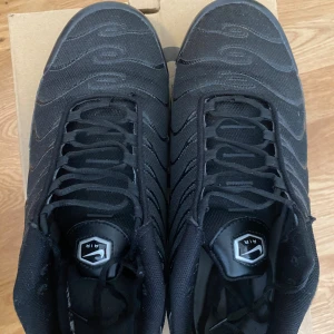 Nike Air Max Plus Tn helsvarta sneakers - Nike Air Max Plus Tn i helsvart med mesh och syntetpaneler, ikoniska vågformade detaljer och Tn Air-logga bak. Skorna har snörning, platt sula och synliga Air Max-enheter i sulan för extra komfort. Perfekt för dig som gillar streetwear och sportig stil. Kontakta mig vid behov av fler bilder!