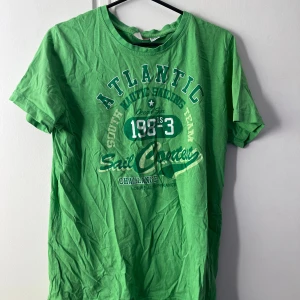 Grön t-shirt från Livergy, M - Fräsch grön t-shirt från Livergy med tryck på bröstet som har texten 'Atlantic Sail Contest 1983'. Klassisk rund hals och korta ärmar. Materialet är mjuk bomull och passformen är normal. Perfekt för chill dagar och enkel att matcha med jeans eller shorts.