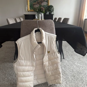 Beige dunväst från Lyle & Scott - Snygg beige dunväst från Lyle & Scott med klassisk gul logga på bröstet. Västen har hög krage, quiltad design och dragkedja framtill. Perfekt för lager-på-lager och enkel att matcha med olika outfits.